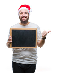 Young caucasian hipster man wearing christmas hat holding blackboard over isolated background pointing and showing with thumb up to the side with happy face smiling