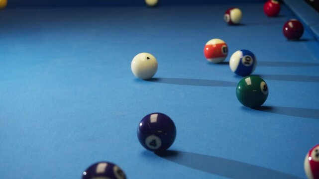 Multicolored American pool balls are scattered on billiard table covered with blue cloth. one of players makes a hit on white ball