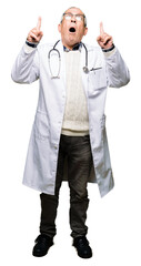 Handsome senior doctor man wearing medical coat amazed and surprised looking up and pointing with fingers and raised arms.