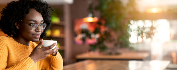 Dreamy young black lady in glasses enjoying cup of coffee in cafe, thinking about future opportunities, close up, copy space