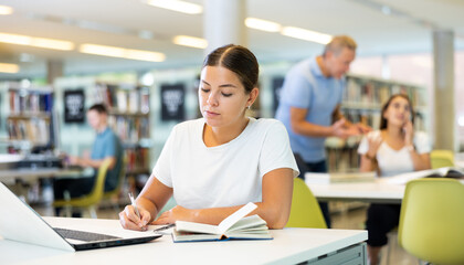 Focused European casual female writing in notepad and using laptop in the library