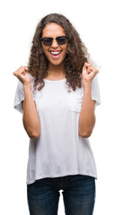 Naklejka premium Young hispanic woman wearing sunglasses excited for success with arms raised celebrating victory smiling. Winner concept.