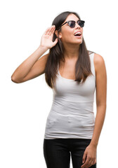 Young beautiful hispanic wearing sunglasses smiling with hand over ear listening an hearing to rumor or gossip. Deafness concept.