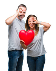 Middle age hispanic casual couple in love holding red heart over isolated background with happy face smiling doing ok sign with hand on eye looking through fingers