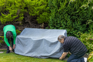 Senior couple put garden seat cover onto outdoor furniture bench set in home backyard garden ready...