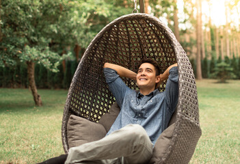 Happy content young man relaxing sitting back in chair smiling in green outdoor nature setting,...