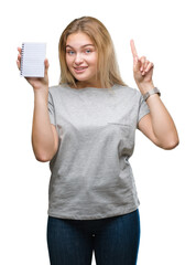 Obraz premium Young caucasian woman holding blank notebook over isolated background surprised with an idea or question pointing finger with happy face, number one