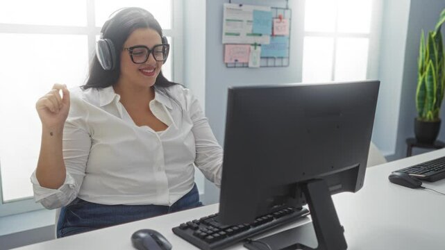 Woman wearing headphones working on a computer in an office while smiling and enjoying music