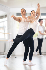 Obraz premium Happy young caucasian man and woman practicing ballet arabesque pose in modern ballroom