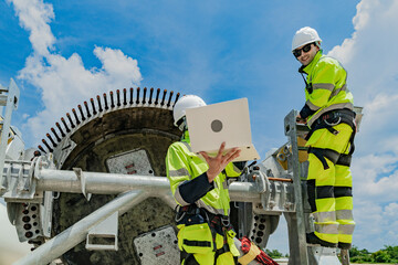 Two engineers work on a wind turbine component, with one using a laptop and the other inspecting parts on a ladder. renewable energy technology, wind power projects, and teamwork in the energy sector.