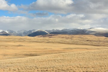 Field roads bending around high hills cross the yellowed steppe at the foot of snow-capped mountains on a sunny autumn day.