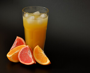 Grapefruit-orange juice with ice on a black background, a tall faceted glass of juice and ripe fruits nearby.