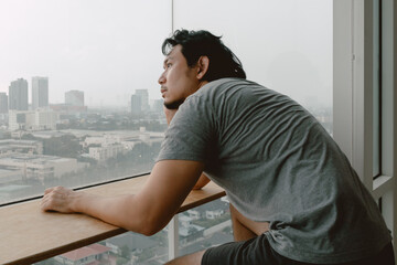 Lonely single asian man looking out to through the windows of apartment.