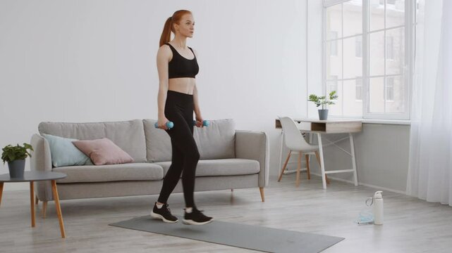 A woman is engaged in a fitness workout in her stylish living room, doing lunges while holding dumbbells. Bright sunlight filters through the window, creating an inviting atmosphere.