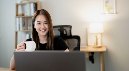 A woman enjoys tea or coffee while working on her laptop in a cozy home office.