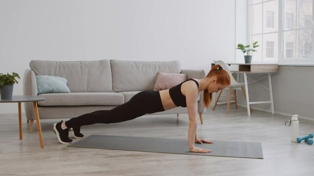 In a modern living room, a young woman in workout attire is completing push-ups on a mat. Natural light filters through the windows, creating an energizing atmosphere for her fitness routine.