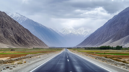 Naklejka premium A straight road leading into the mountains cuts through a wide valley between majestic snow-capped mountain ranges. In the distance, a cloudy sky looms on the horizon