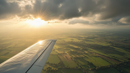 View from an airplane window showing the wing flying over green fields at sunset. Sun rays break through the clouds, illuminating the horizons and highlighting the beauty of the landscape