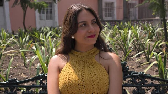 Mujer feliz mexicana y latina est&aacute; sentada en la banca de un parque y sonr&iacute;e