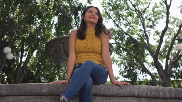 Mujer Feliz Mexicana y latina sentada en un parque, delante de una fuente, sonriendo y viendo pasar a la gente.
