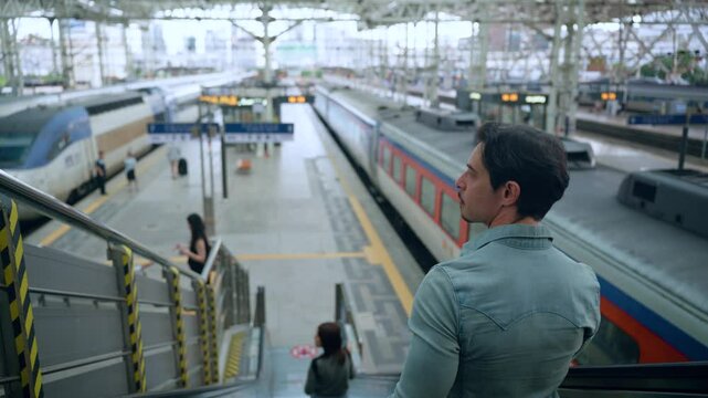 Australian Man Traveler Riding Escalator Down to Platforms of Seoul Train Station, View Of Railway Tracks and Locomotives - over shoulder view