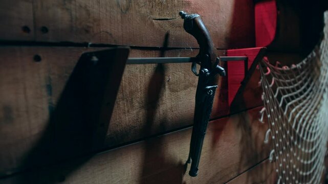 An antique flintlock pistol is mounted on a rustic wooden wall beside a section of fishing net. Vintage firearm and nautical decor of a maritime pirate ship