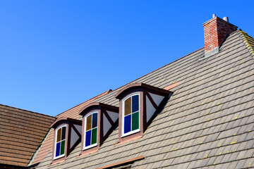 A sloped tiled roof with three dormer colorful stained glass windows, and a brick chimney, set...