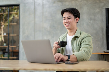 A handsome Asian man is working remotely from a coffee shop, enjoying coffee while working on laptop