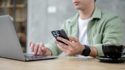 A man holding his phone while typing on his laptop keyboard, working remotely from a coffee shop.