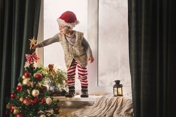 boy 3-4 years old in red and white stripped pajamas and Santa Claus hat plays on the window sill near the decorated New Year tree with lanterns and gingerbread house. Christmas symbols, holiday card	