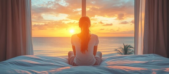 Woman sitting on a bed facing a window with ocean sunset view.