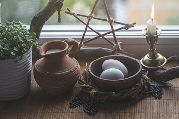 Magic home set. Atmospheric altar on windowsill with eggs, pentacle, the first greens in the pot, candle, birds feathers and clay jug with milk, selected focus.