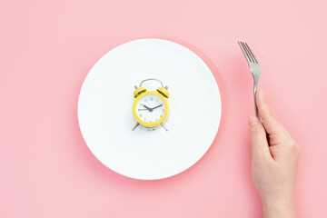 Female hand holding a fork next to an empty plate with alarm clock on a pink background close-up...