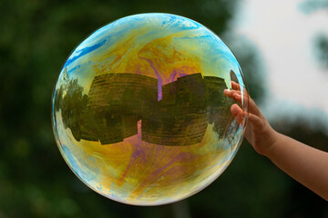 A child plays with soap bubbles. A child's hand touches a soap bubble. There is a reflection of a building in the balloon, and the balloon resembles the Earth.