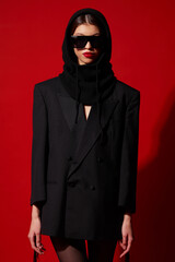 Fashion asian female model in black jacket, balaclava, trendy sunglasses. Red lips, red background.