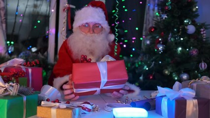Santa Claus with holiday gift boxes wrapped in color paper at table in workshop next to many presents and Christmas tree, festive invitation.
