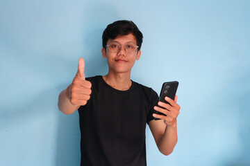 young Asian man in black t-shirt holding mobile phone and making thumb up gesture, approve good thing