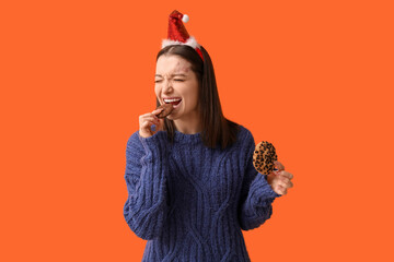 Beautiful young woman in Christmas hat eating tasty cookies on orange background