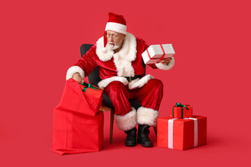 Santa Claus with many gift boxes and bag sitting on armchair against red background