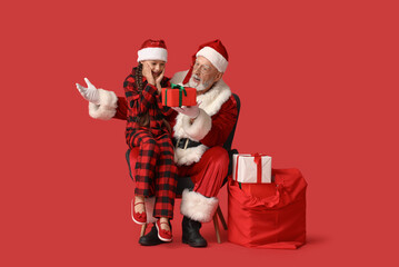 Shocked Santa Claus and cute little girl with gift boxes sitting on armchair against red background