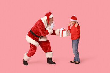 Santa Claus and cute little girl with gift box showing silent gesture on pink background
