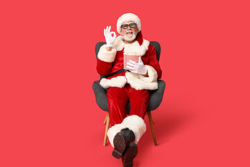 Santa Claus in 3D glasses with bucket of tasty popcorn showing ok gesture on red background