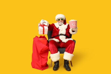 Santa Claus holding bucket of tasty popcorn and bag with gifts on yellow background