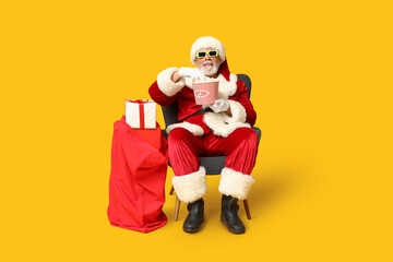 Santa Claus holding bucket of tasty popcorn and bag with gifts on yellow background