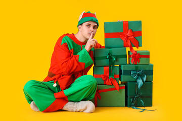 Handsome young man in elf's costume with gift boxes showing silent gesture on yellow background
