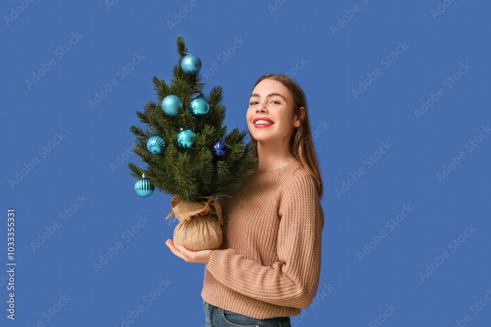 Wall mural Beautiful young woman holding Christmas tree with balls on blue background