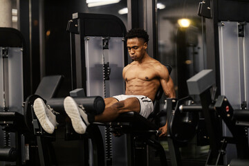 Powerful tough shirtless black sportsman doing workouts on leg extensions machine.