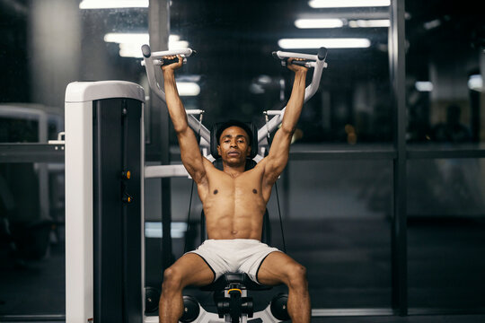 Front view of active strong muscular diverse bodybuilder doing exercises on shoulder press machine at gym. - Powered by Adobe