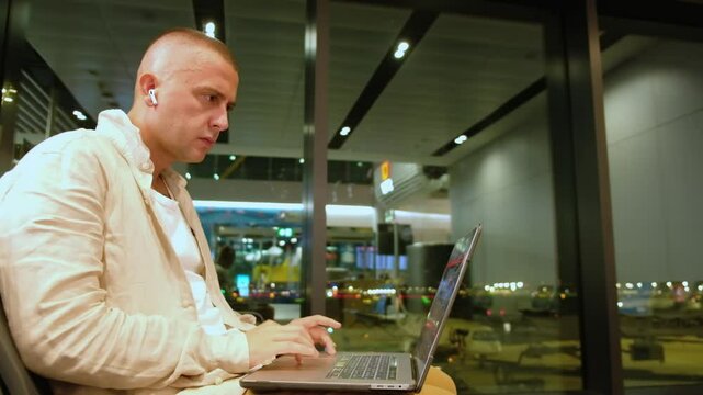 Young adult caucasian man woks on laptop at night airport lounge. Solving problems on-the-go concept. Shocked man doomscrolling during layover at airport
