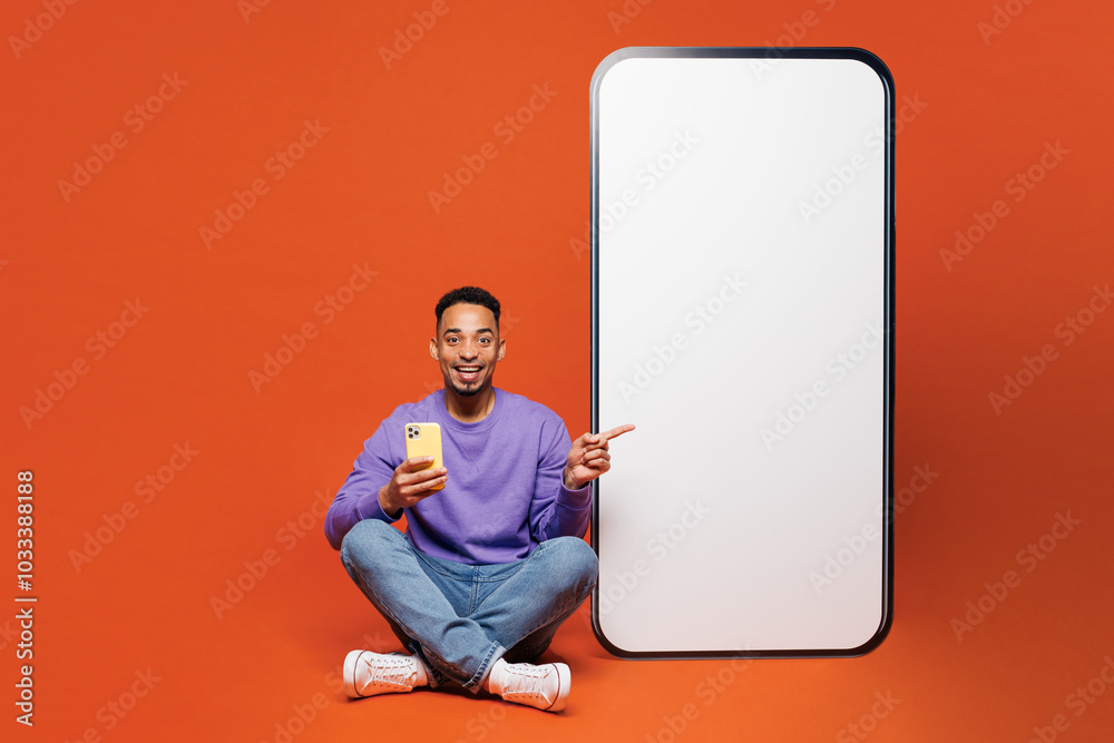 Poster Full body happy young man wear purple sweatshirt casual clothes sits point on big huge blank screen mobile cell phone smartphone with area isolated on plain red orange background. Lifestyle concept.